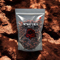 Eine durchsichtige Kunststoffverpackung mit dem Aufdruck 'M'NATURA BAITS' und einem roten Fischlogo mit der Aufschrift 'MK SPICE' steht auf erdigem, rotbraunem Untergrund. Die Verpackung ist mit zahlreichen feurig rot gefärbten, runden Boilies gefüllt. Ein kleiner Hacken mit Boilie Kugel ist oben rechts auf der Verpackung abgebildet. Die Produktbezeichnung 'Boilies feurig rot würzig' ist durch das Kontextwissen gegeben.